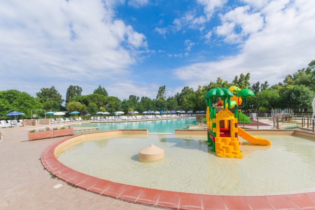 Piscina con un'area giochi d'acqua colorata e ombrelloni bianchi sullo sfondo.