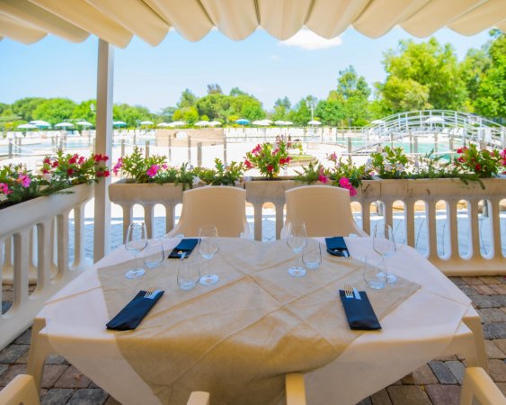 Tavolo apparecchiato su terrazza con vista su piscina e fiori colorati attorno.