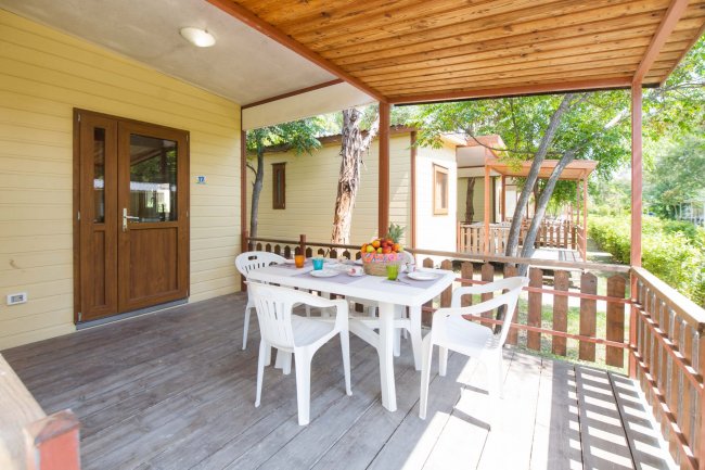 Veranda con tavolo bianco, sedie e recinzione in legno, circondata da alberi.