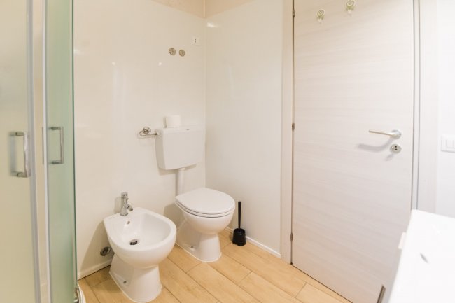 Bagno con bidet, water, scovolino e porta bianca chiusa, pavimento in legno chiaro.