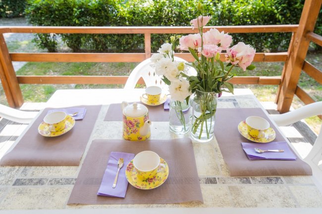 Tavolo apparecchiato con tazze floreali e vaso di fiori su un terrazzo.