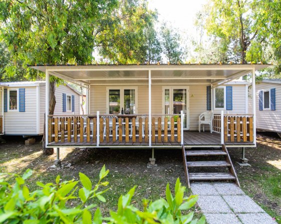 Casa mobile con veranda, staccionata in legno, tavoli e sedie, circondata da alberi.