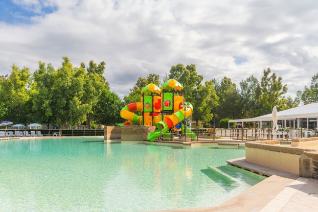 Piscina con scivoli colorati al centro, circondata da alberi e panchine.