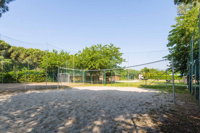 Campo da beach volley con rete e sabbia, circondato da alberi e una recinzione.