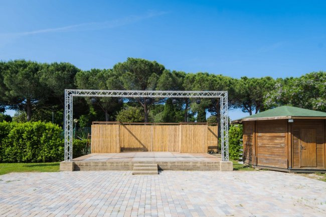 Palco all'aperto con struttura metallica, pavimento in mattoni e capanno vicino.