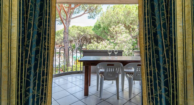 Tavolo e sedie su un balcone, viste su alberi e natura attraverso tende aperte.