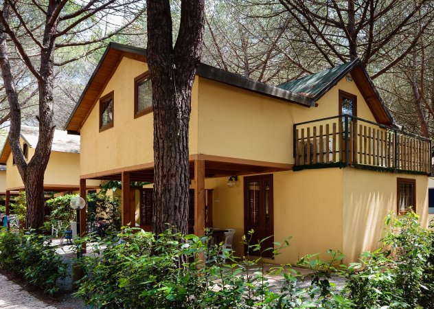 Una casetta gialla a due piani con balcone, circondata da alberi e vegetazione.
