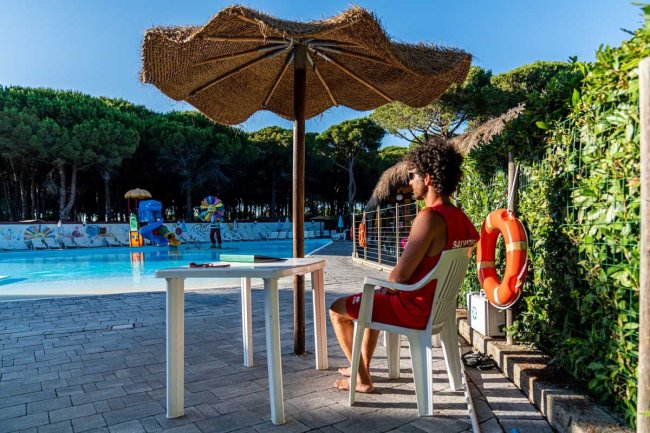 Uomo seduto sotto un ombrellone di paglia vicino a una piscina; alcuni bambini giocano in acqua.