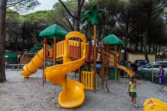 Parco giochi con scivoli gialli e verdi. Bambini che giocano nella sabbia, alberi intorno.