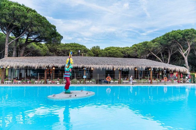 Piscina azzurra con statua di surfista, tetto di paglia e alberi sullo sfondo.