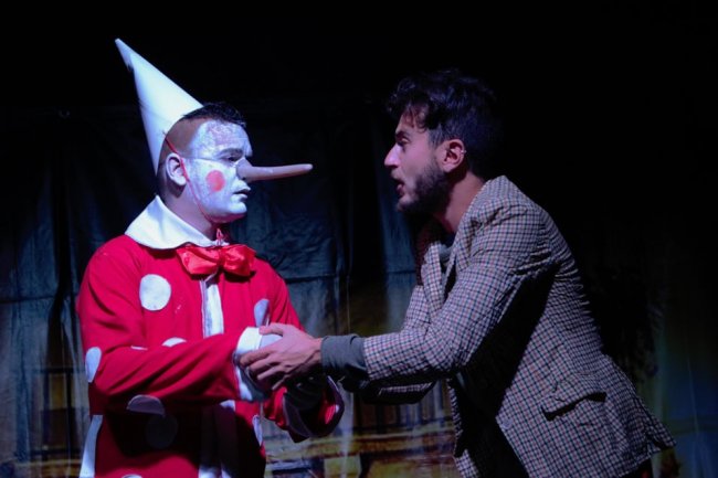 Un uomo si esibisce con un costume da clown con lungo naso mentre parla con un altro uomo.
