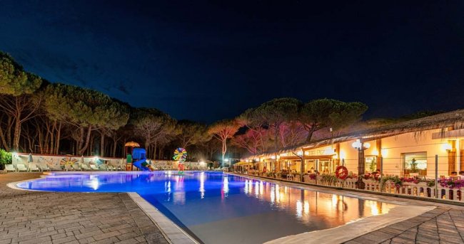 Piscina illuminata di notte con area ricreativa e pini sullo sfondo.