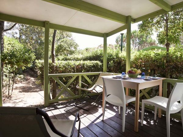 Veranda in legno con tavolo, sedie bianche e vegetazione circostante.