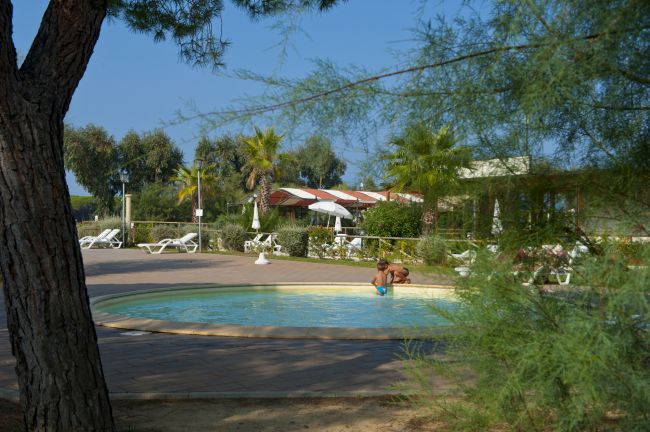 Piscina rotonda tra alberi con un bambino che gioca in acqua e lettini in lontananza.