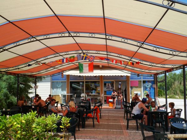 Area esterna di un bar con copertura e sedie nere. Persone sedute ai tavoli e bandiere appese.