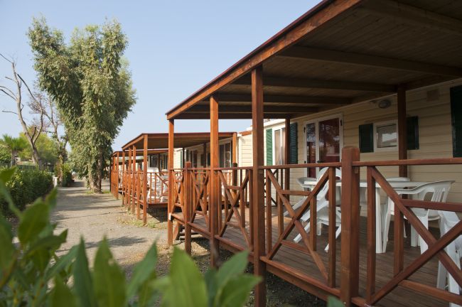 Bungalow con veranda in legno e sedie bianche accanto a un sentiero alberato.