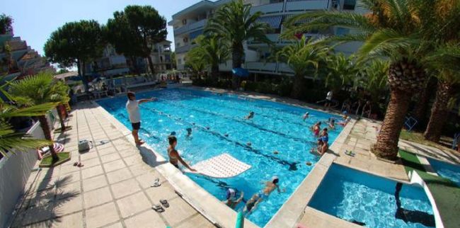 Piscina affollata con persone che nuotano, circondata da palme e edifici residenziali.