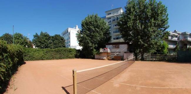 Campo da tennis all'aperto con rete, contornato da alberi e edifici bianchi.