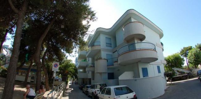 Edificio moderno angolare con balconi curvi, strada con auto parcheggiate e alberi.