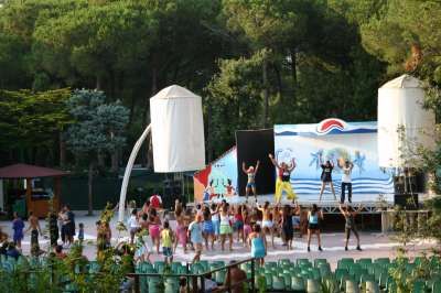 Persone fanno esercizi su un palco all'aperto, circondate da sedie e alberi.