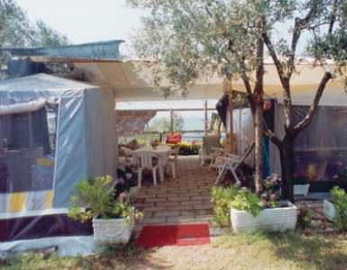 Una veranda coperta da una tenda, con tavoli, sedie e piante in vasi, situata tra alberi.