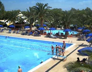 Piscina all'aperto con ombrelloni blu e palme intorno. Persone sulla piattaforma e sdraio.