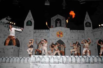 Spettacolo teatrale con performer in abiti bianchi e un edificio di scenografia sullo sfondo.