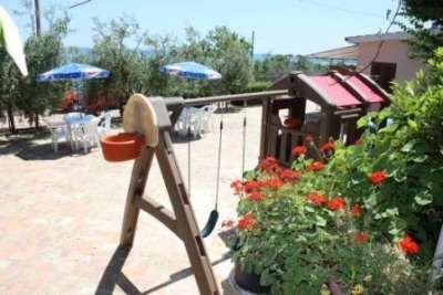 Giardino con altalena di legno, fiori rossi e tavoli con ombrelloni sullo sfondo.