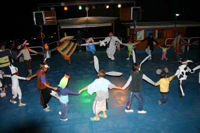Bambini in cerchio, mascherati, danzano su un palco all'aperto illuminato.