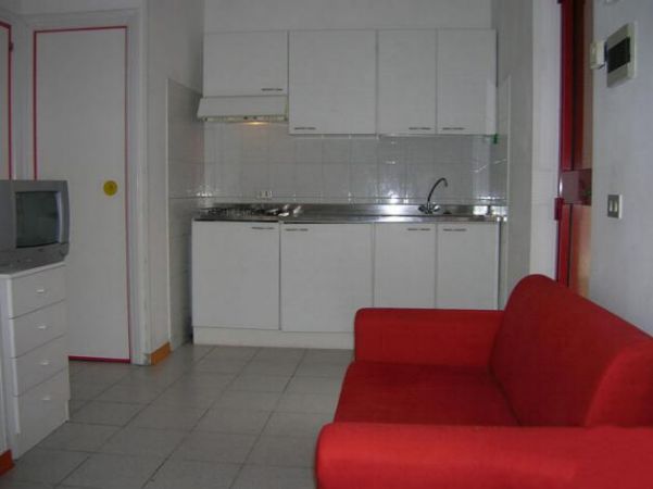 Un soggiorno con un divano rosso, cucina a vista e TV su un mobile bianco.