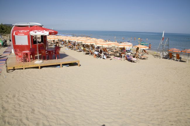 Spiaggia sabbiosa con lettini, ombrelloni arancioni e chiosco rosso.