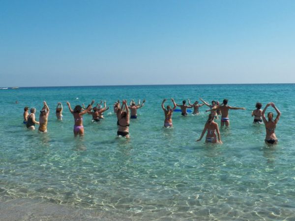 Persone fanno esercizi in mare con le braccia alzate sotto un cielo azzurro.