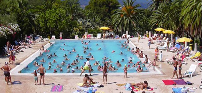 Ampia piscina esterna affollata di persone, con lettini e palme sullo sfondo.