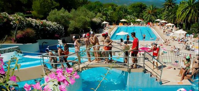 Persone su un ponte sopra una piscina con scivoli, circondata da fiori e verde.