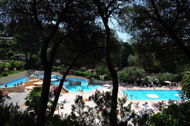 Piscina affollata tra gli alberi con persone su sdraio.