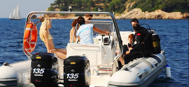 Persone a bordo di un gommone con due motori in un mare calmo, costa rocciosa in lontananza.