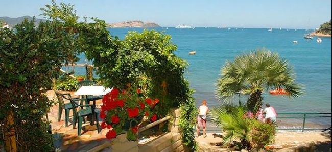 Terrazza sul mare con tavolo e sedie, fiori rossi e vista su una spiaggia e mare.