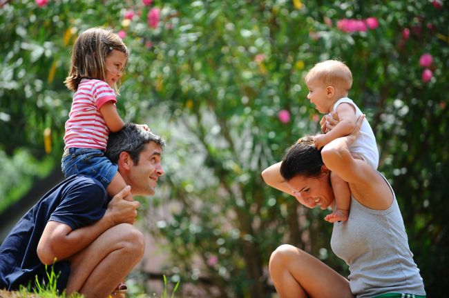 Bambini sorridenti sulle spalle di due adulti in un giardino fiorito.