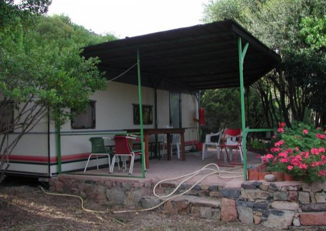 Patio coperto con tavolo e sedie, accanto a una roulotte tra gli alberi.
