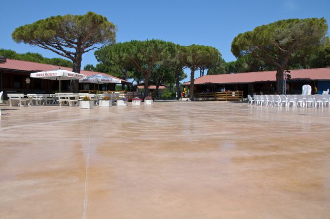 Spiazzo in cemento con sedie e tavoli, circondato da alberi e edifici bassi.