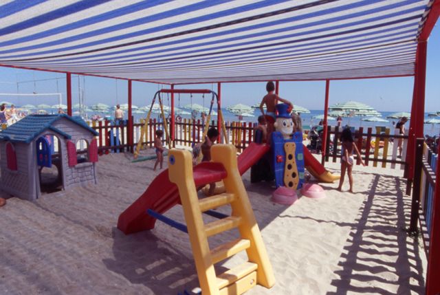Area giochi sulla spiaggia con altalene e scivoli sotto una tettoia a righe blu.