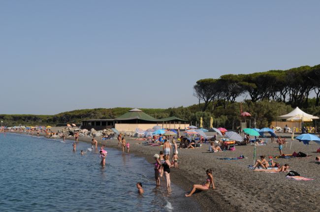 Spiaggia affollata con persone in costume, ombrelloni colorati e una struttura in lontananza.