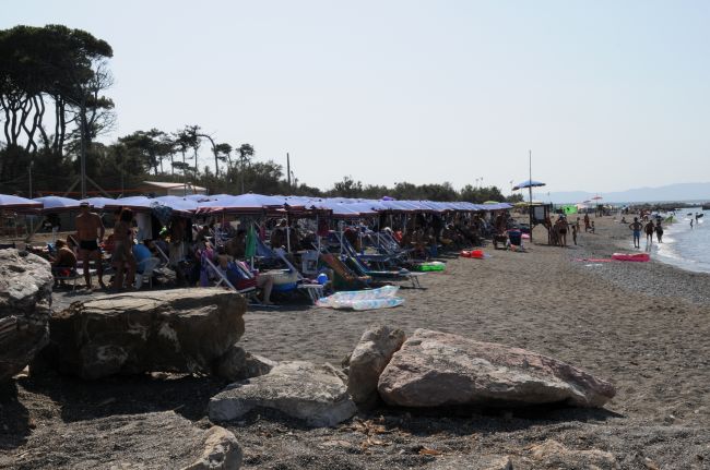 Spiaggia affollata con ombrelloni e lettini, vicino al mare con grandi rocce.