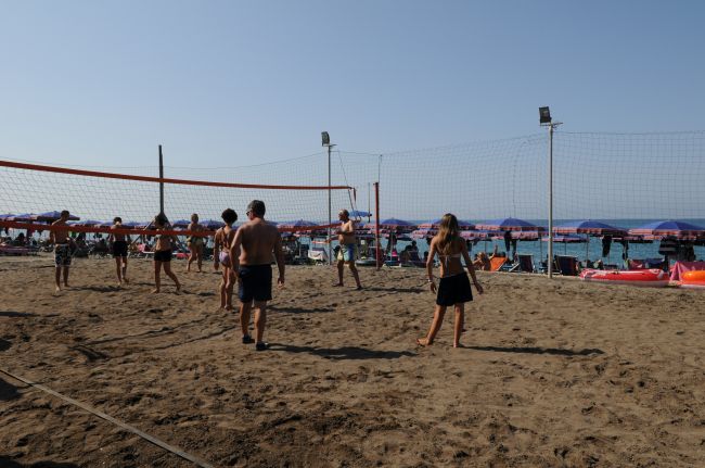 Partita di beach volley sulla sabbia vicino al mare, con ombrelloni sullo sfondo.