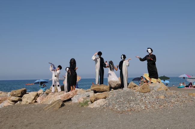 Persone in costumi teatrali neri e bianchi su una spiaggia rocciosa con il mare sullo sfondo.