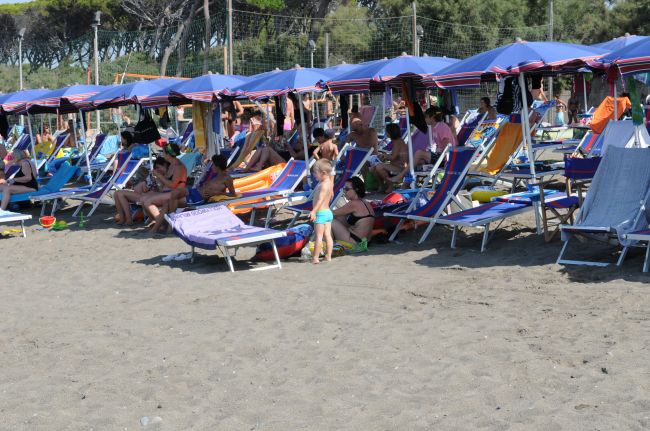 Persone sdraiate su lettini sotto ombrelloni blu in spiaggia, con un bambino che gioca.