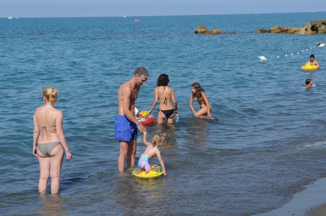 Persone in acqua lungo una spiaggia, tra cui un uomo e un bambino con un salvagente.