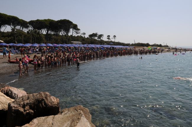 Una spiaggia affollata con persone in fila lungo la riva, ombrelloni blu e alberi sullo sfondo.