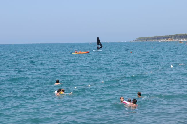 Bagnanti nel mare, kayak arancione e vela nera sullo sfondo.