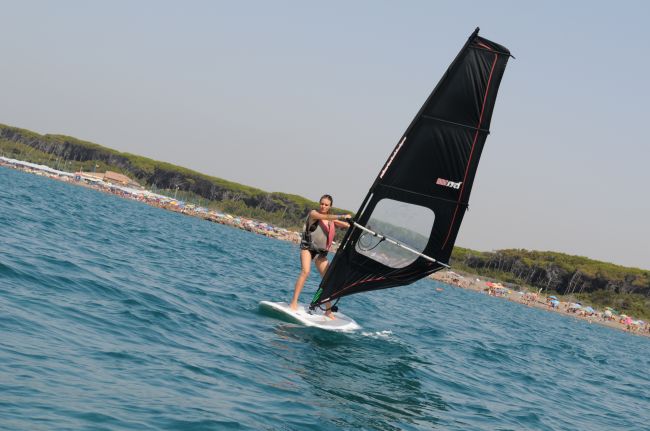 Persona che fa windsurf su acqua azzurra, vicino a una spiaggia affollata con pineta.